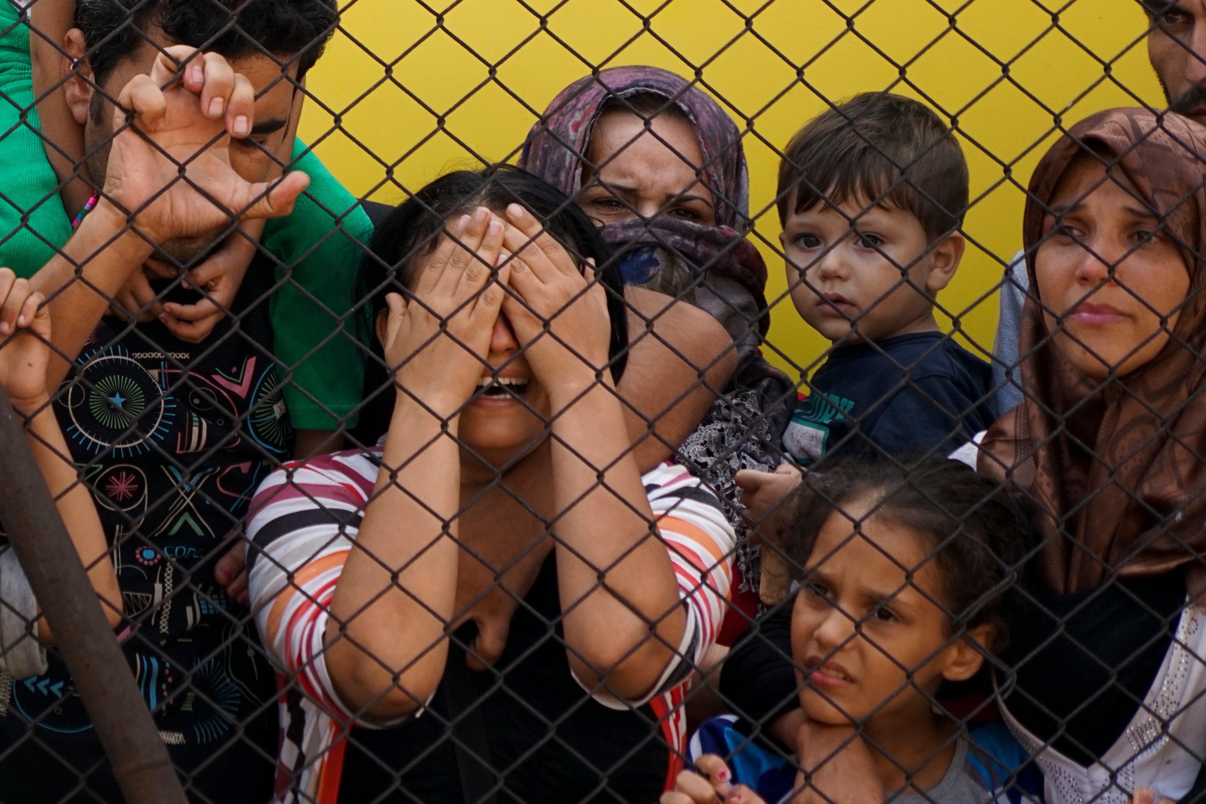 A group of children, women and a young man behind a chain link fence, one woman with hands over her eyes, wailing