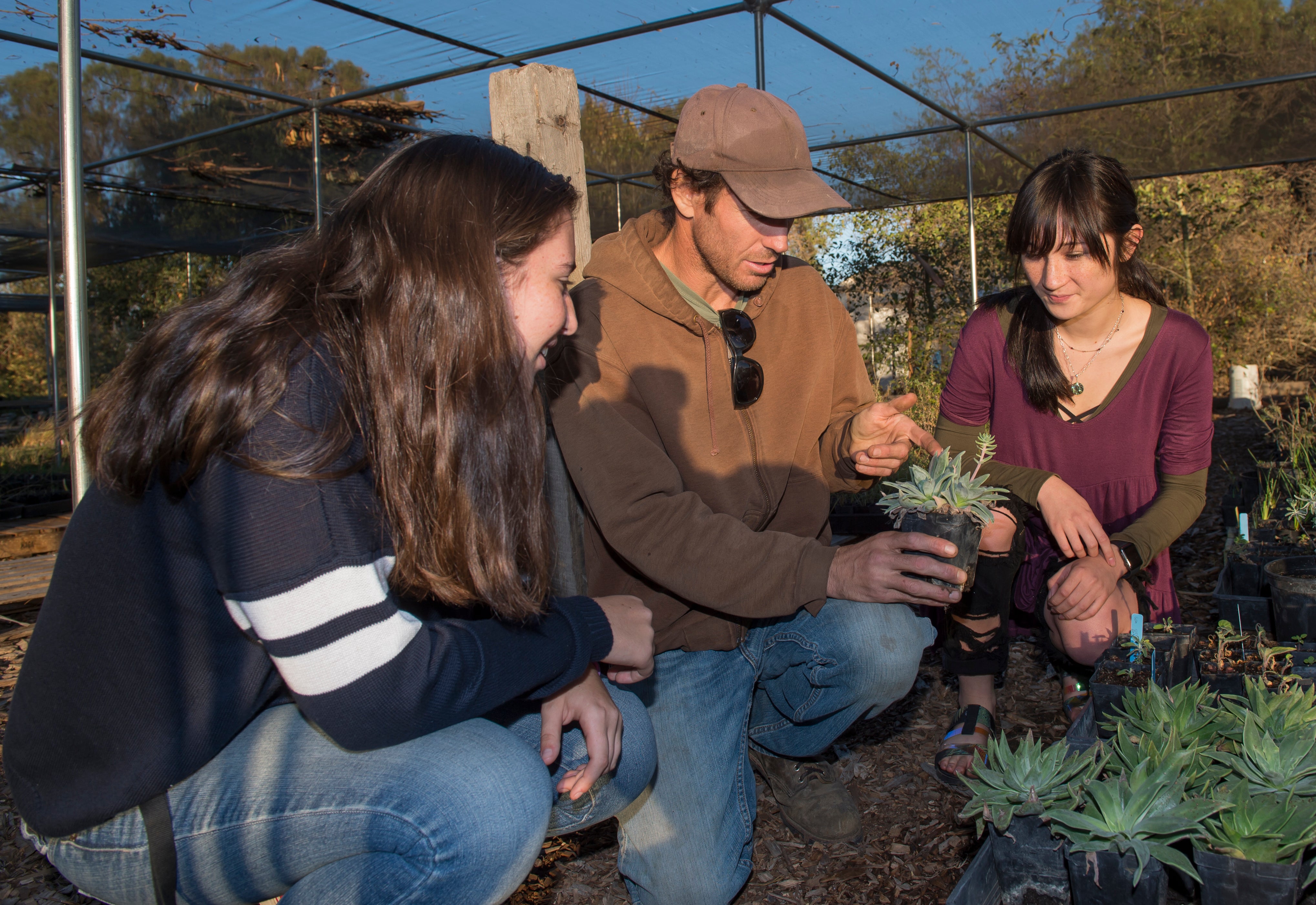 Two young women watching a professor in a brown ballcap with a plant, in a garden
