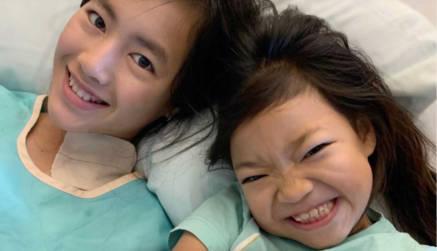 Ada and Lily Finlayson, school-aged, take a selfie wearing green medical gowns.