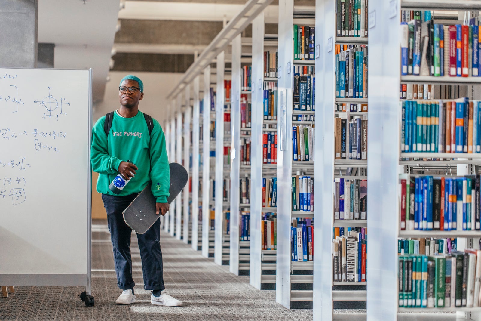 Young man wearing green sweatshirt carrying a skateboard leans against a white board next to library shelves
