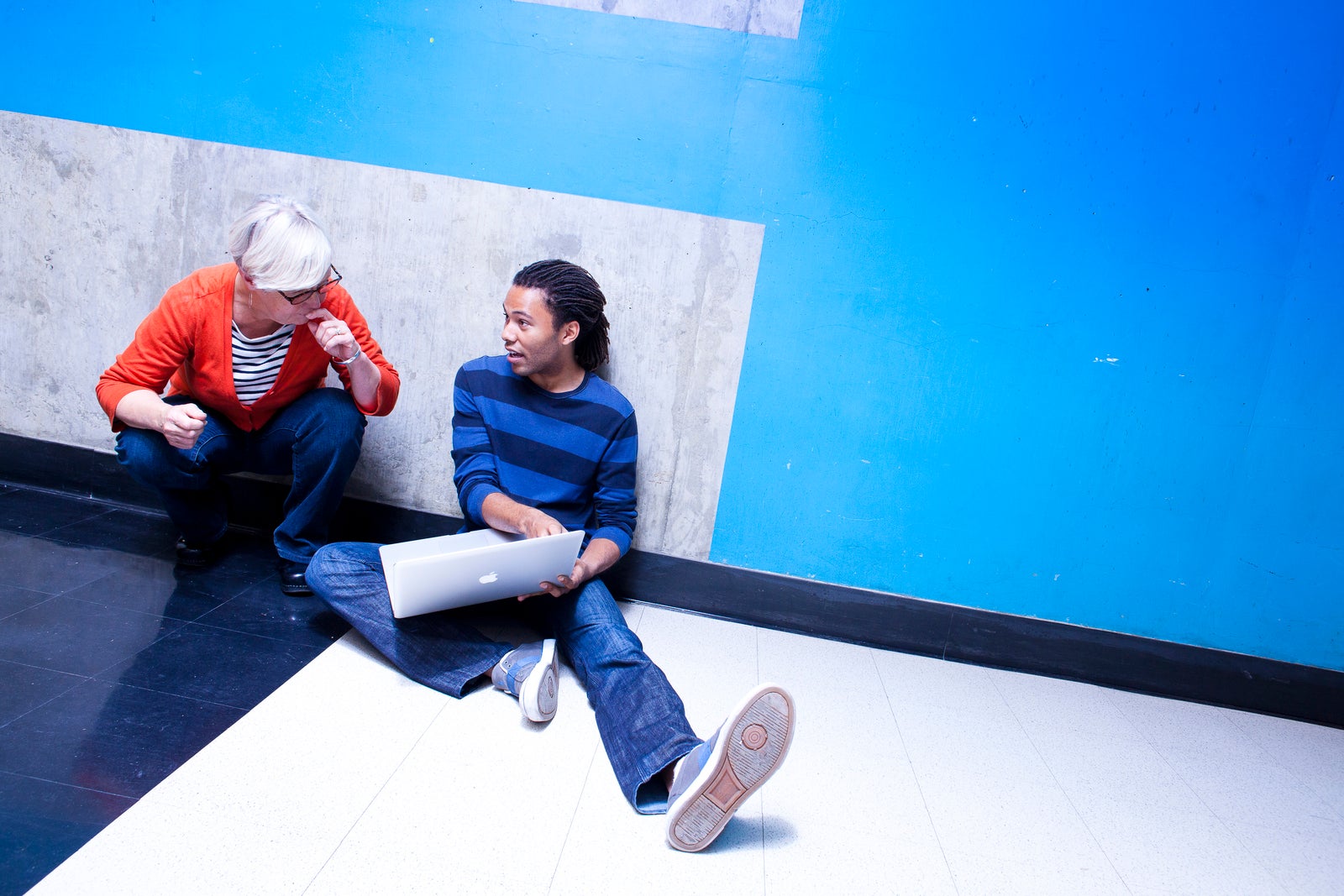 A female professor crouches next to a male student, who's sitting with his legs stretched out in a hallway with a blue wall, pointing to something on a Mac laptop screen