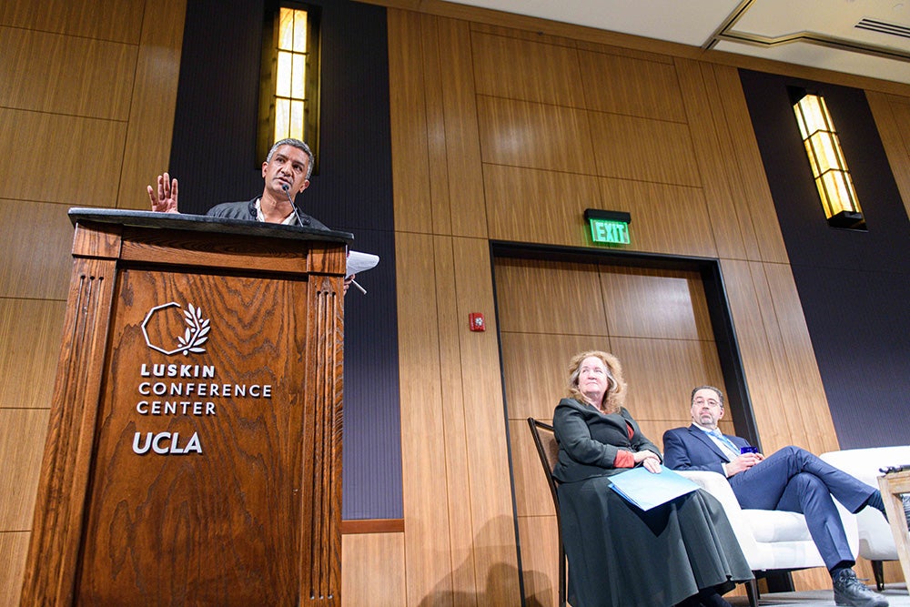 Ramesh Srinivasan gesticulates at a podium branded with UCLA Luskin Center. UC Provost Katherine Newman and MIT Professor Daron Acemoglu sit on stage next to the podium.