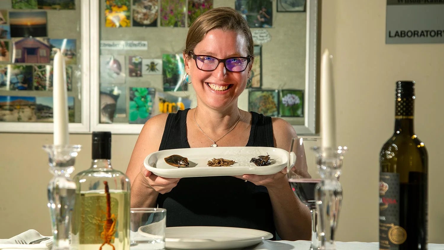 A grinning woman with glasses holds up a plate of bugs at a dinner table with olive oil and wine nearby