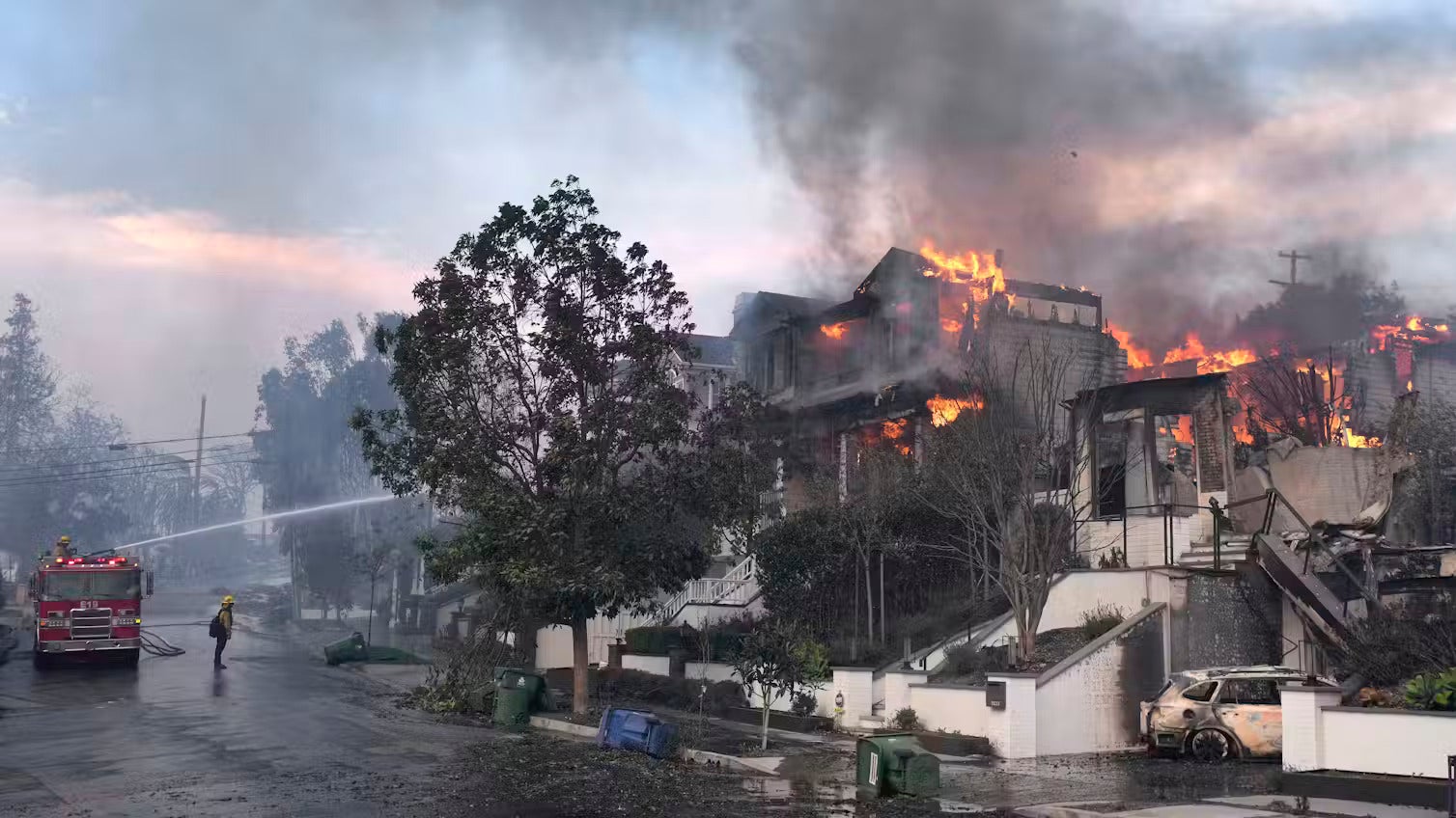 The upper levels of a home on fire in a nice neighborhood, a fire truck spraying the home with water from a hose while a firefighter looks on
