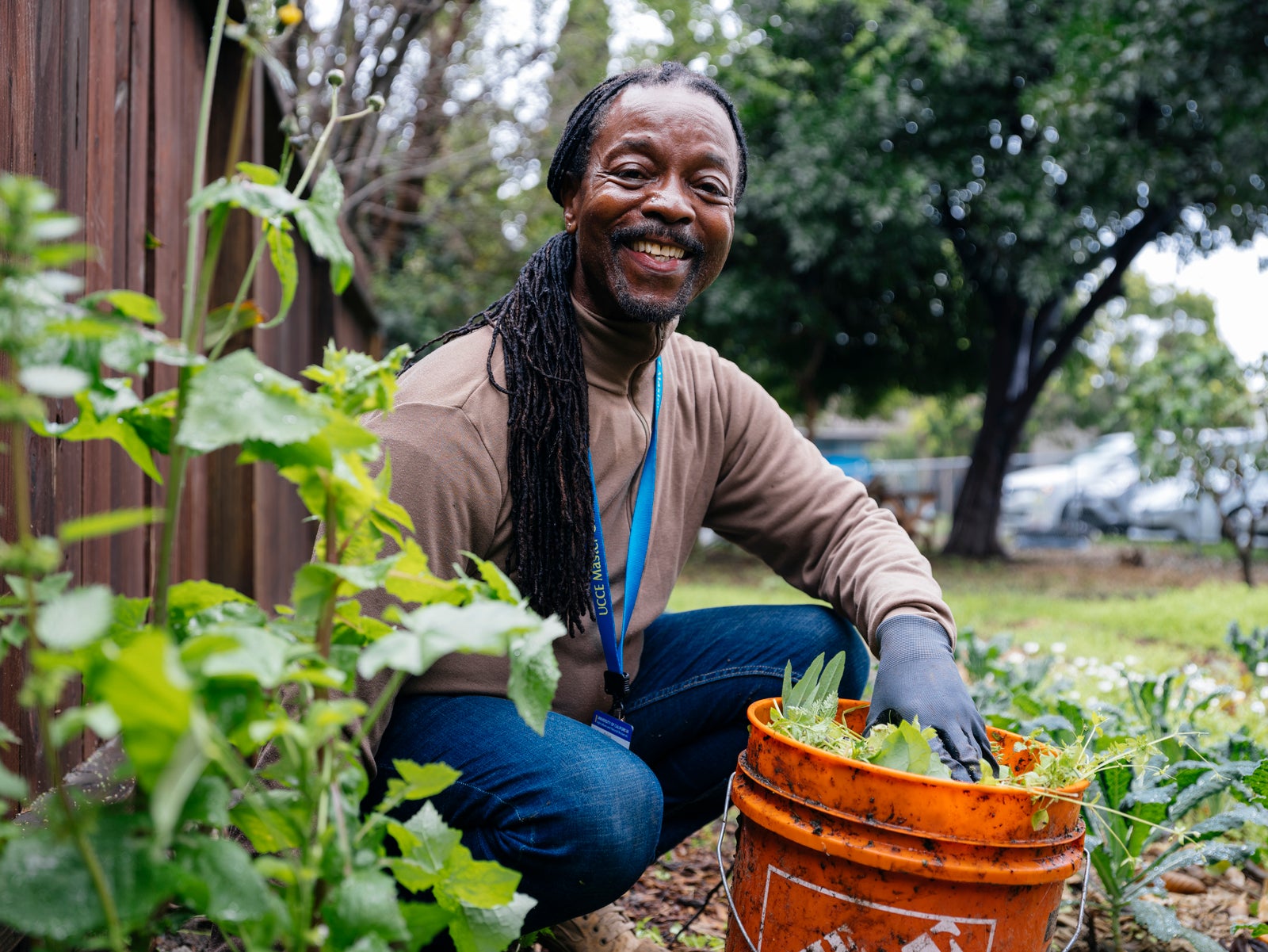 A man with long braids pauses from weeding a garden to smile for a photo