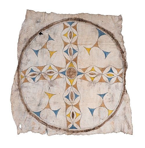 A Tikuna/Magüta item with a pattern of golds, browns, and blues in a cross-like shape, surrounded by a ring, with four three-pronged shapes in the space between right angles and the ring within