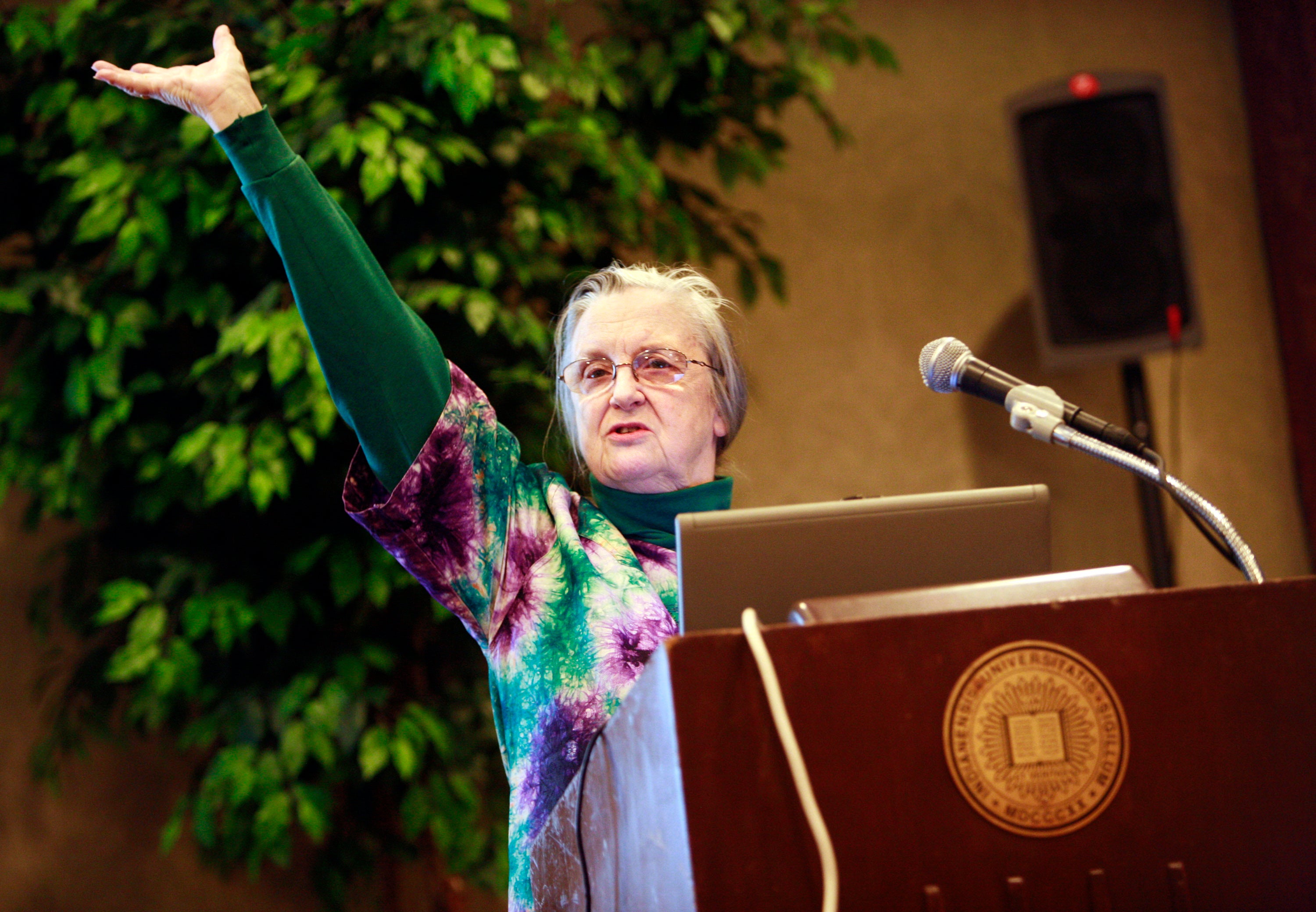 A woman stands before a podium, gesturing grandly with her arm in the air