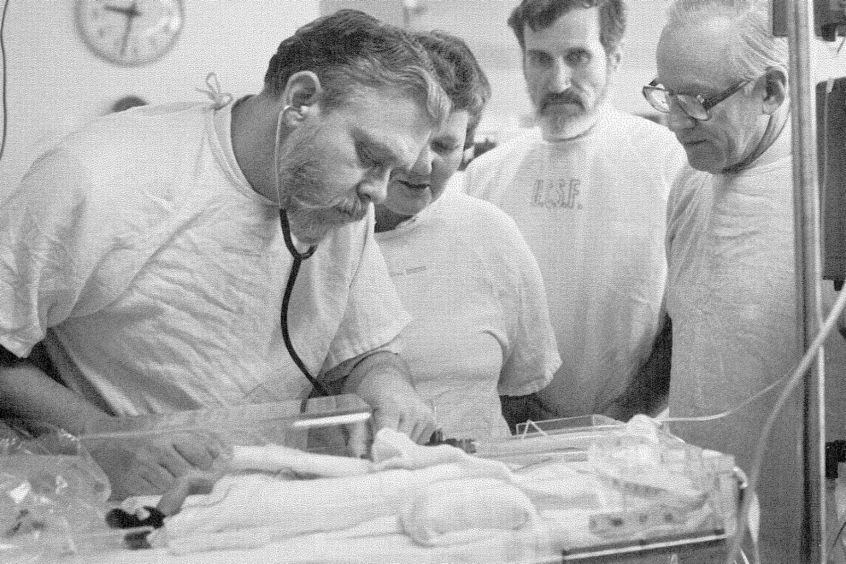 Four doctors examine a small baby in an incubator in a black-and-white photo