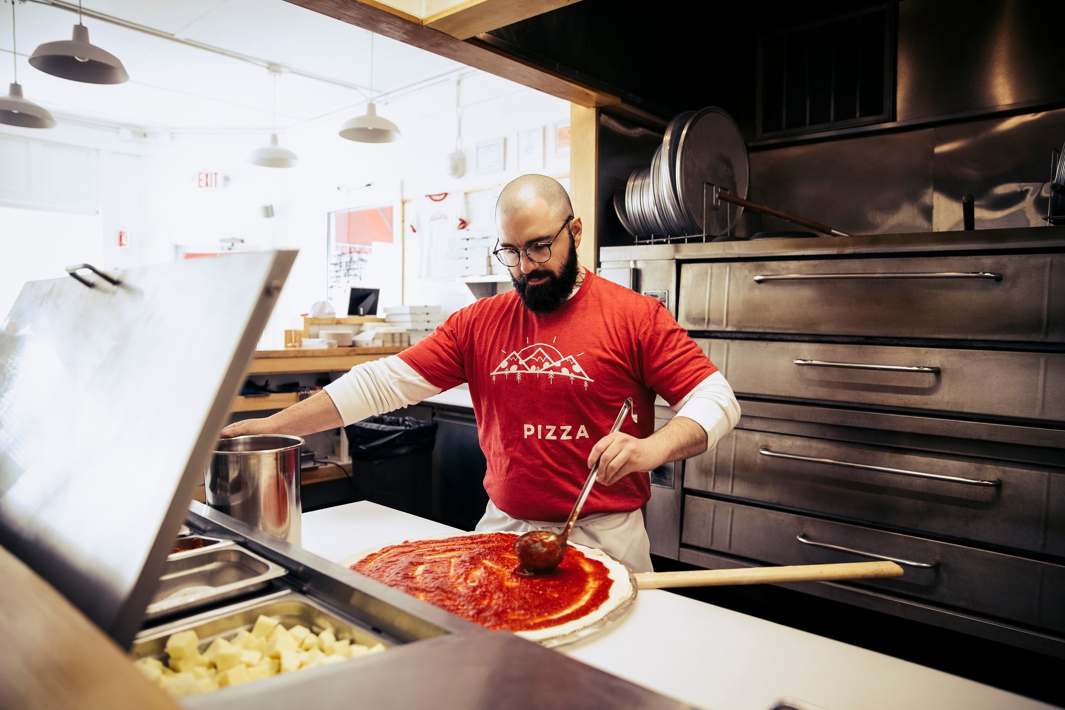 A chef puts pizza sauce on pizza dough in a commercial kitchen