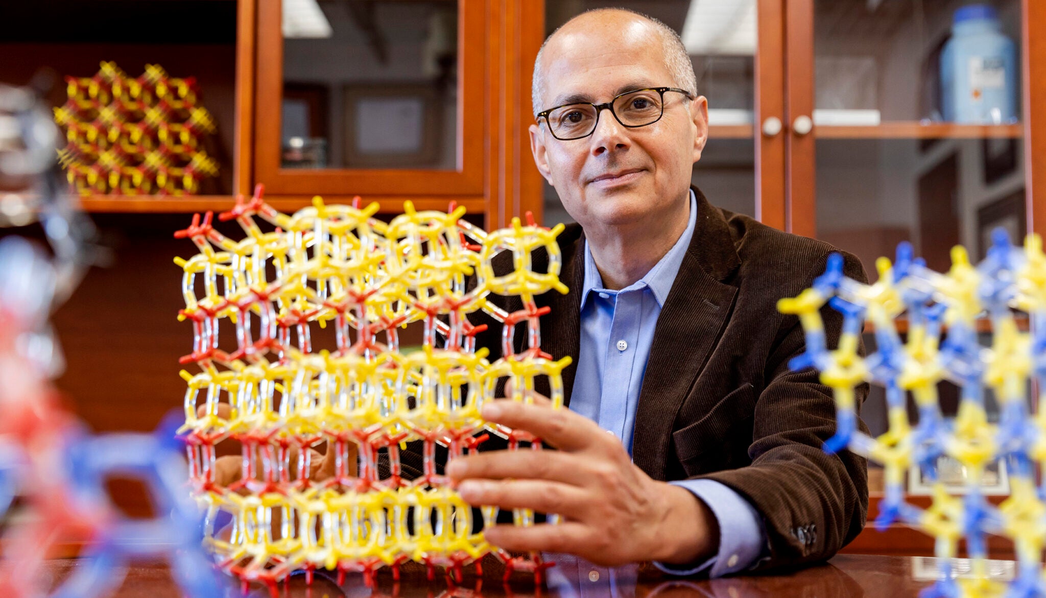A man with glasses and a brown jacket holds molecular models on a table