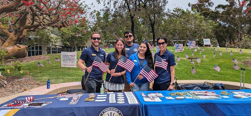 A group of five people wearing blue standing behind a table holding small American flags, with many more flags planted in the lawn behind them