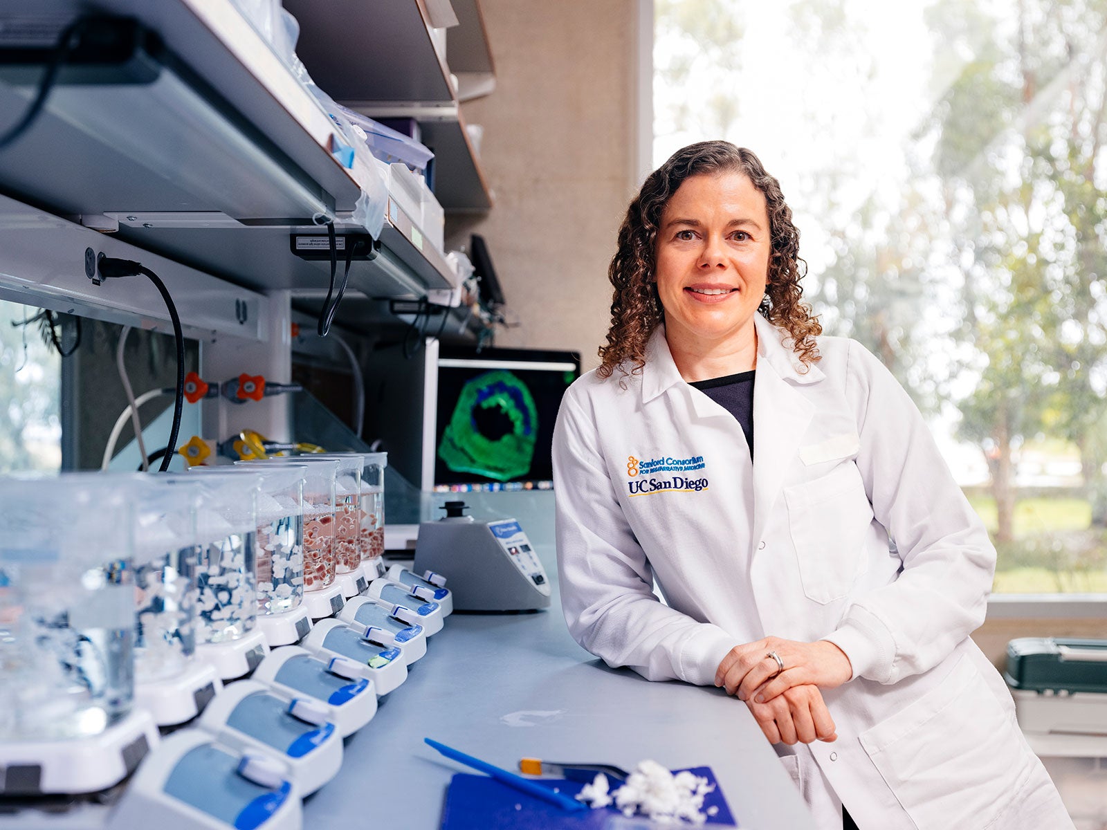 Karen Christman, wearing a white lab coat with UCSD embroidered on it, leans against a lab bench with beakers on bunson burners, smiling at the camera. 