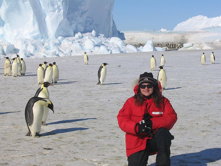 A woman in a red jacket kneeling in Antarctica with a number of penguins behind her