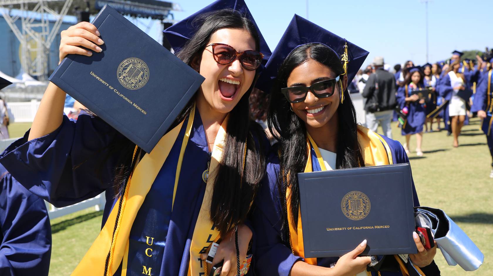 Smiling students at graduation wearing caps and gowns and holding UC Merced degrees