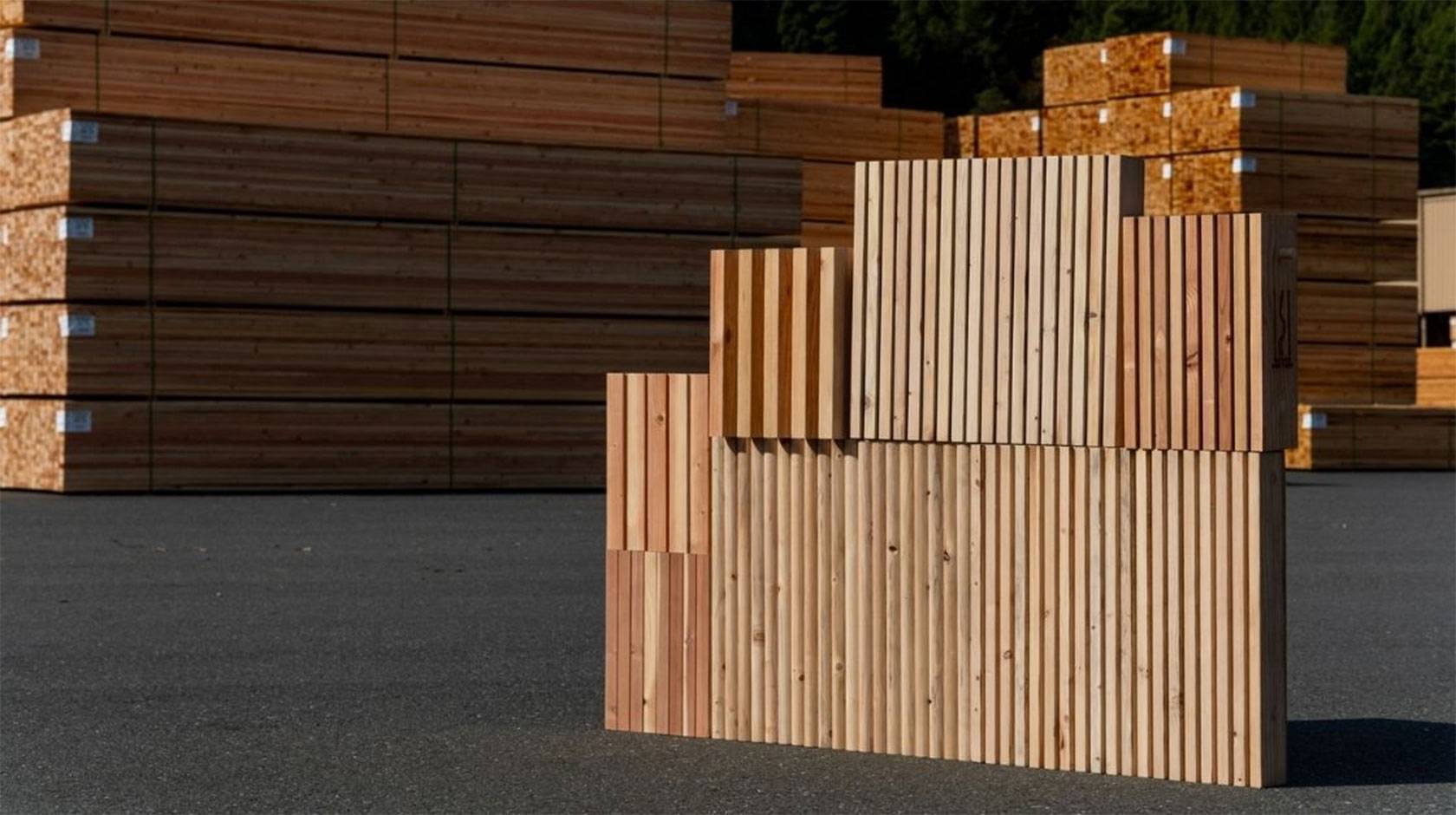 Stacked wooden panels with vertical slats arranged in stepped blocks on an outdoor asphalt surface, with large piles of lumber stacked in the background at what appears to be a timber yard.