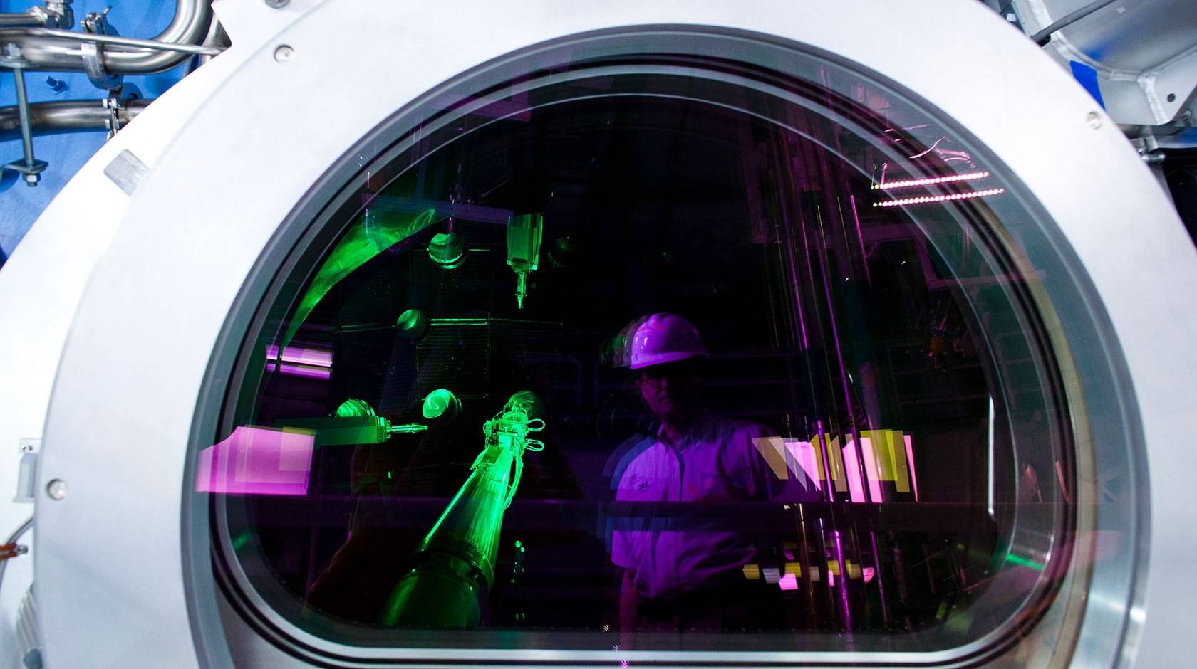 A person wearing a white hard hat is reflected in the circular viewing window of a large scientific instrument, while bright green laser beams and purple lighting illuminate the interior of a laboratory filled with optical equipment and metal piping.