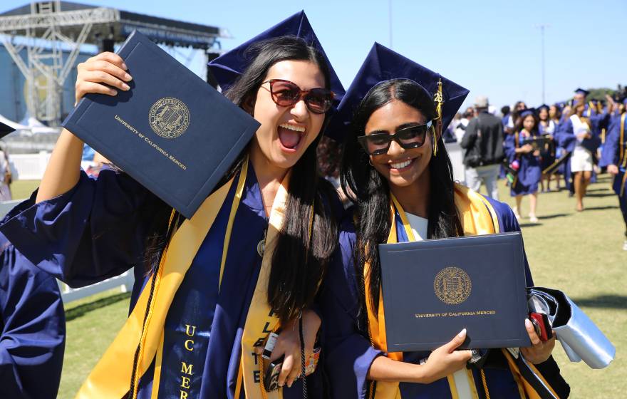 Smiling students at graduation wearing caps and gowns and holding UC Merced degrees