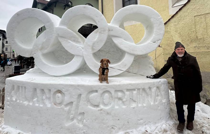 In front of a snow sculpture of the Olympic rings that reads Milano Cortina, a man stands next to a service dog sitting on the sculpture
