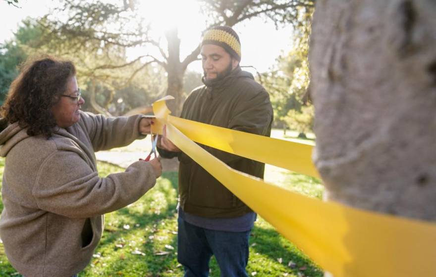 A woman cuts a long yellow ribbon tied to a tree a man is holding