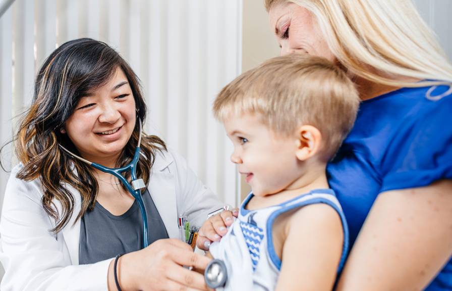 A smiling female doctor uses a stethoscope on a happy toddler sitting on a mother's lap. 