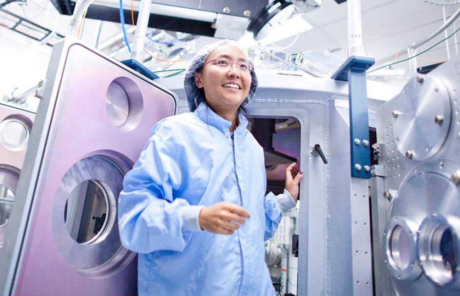 A person in a blue lab coat and hair net stands inside a laboratory, surrounded by large scientific equipment.
