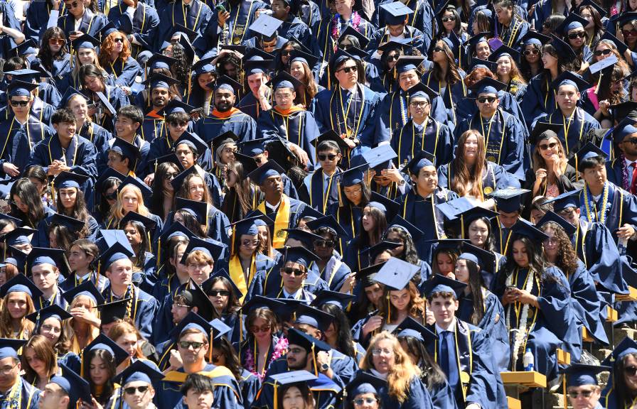 A high-angle shot of a dense crowd of university graduates in blue caps and gowns filling stadium bleachers, accented by colorful leis, stoles and sunglasses under bright sunlight.