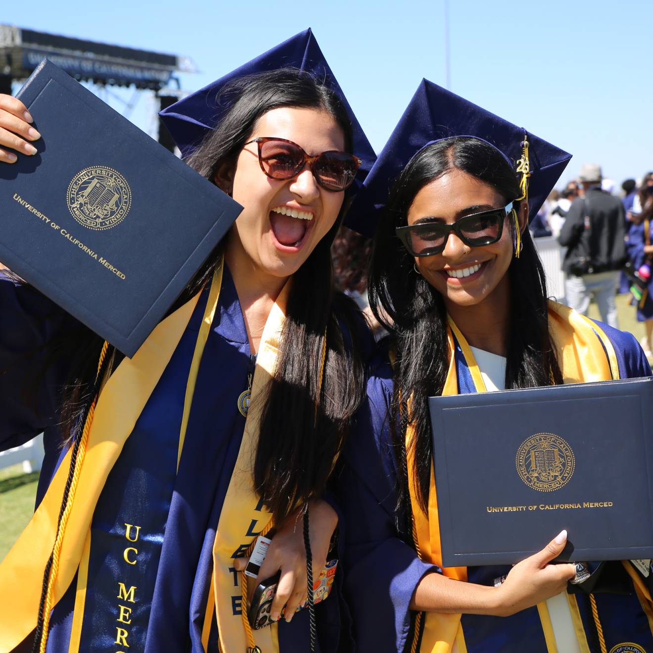Smiling students at graduation wearing caps and gowns and holding UC Merced degrees